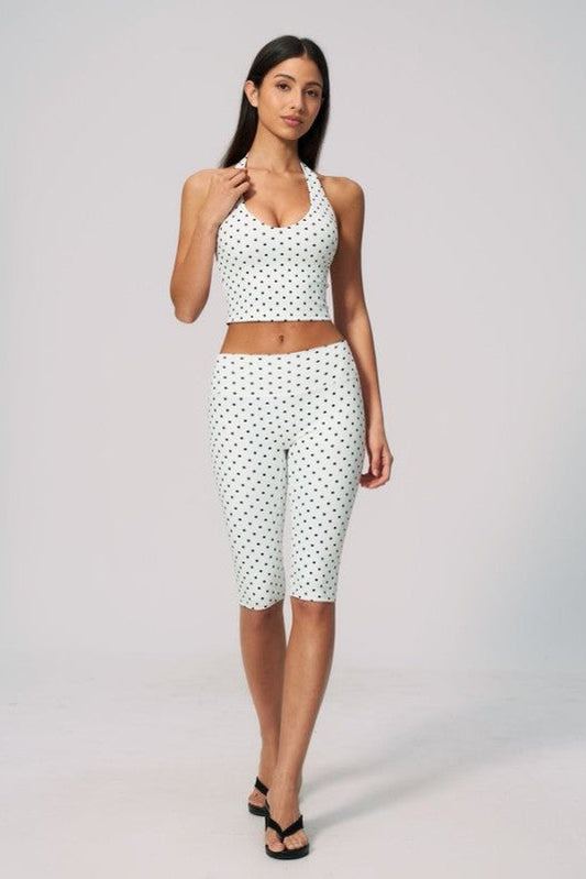 A woman modeling a matching two-piece activewear set consisting of a white halter crop top and white capri leggings with a small black polka dot pattern, set against a plain light gray studio background.