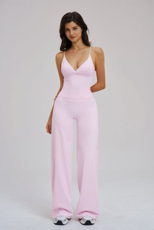 A woman modeling a light pink sleeveless jumpsuit with a deep V-neckline and wide-leg pants, standing against a plain light gray studio background.