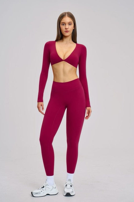 A woman modeling a matching deep magenta long-sleeve crop top and high-waisted leggings set, paired with white socks and white sneakers, against a solid light gray studio background.