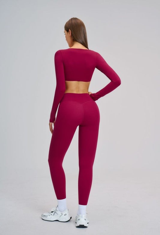 A woman is shown from the back wearing a matching magenta long-sleeve crop top and high-waisted leggings set, paired with white socks and white sneakers, against a light gray studio background.