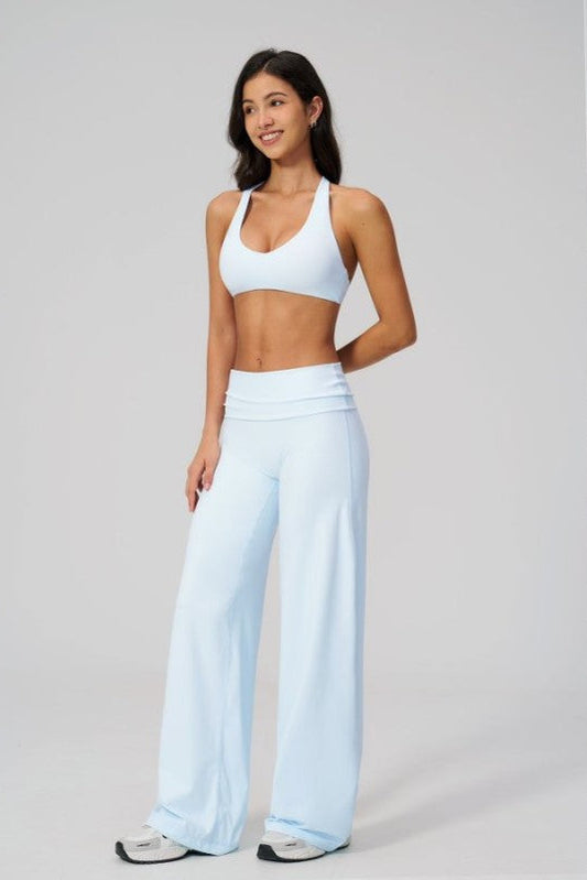 A woman modeling a light blue activewear set consisting of a sports bra and high-waisted wide-leg pants, photographed against a neutral light gray studio background.